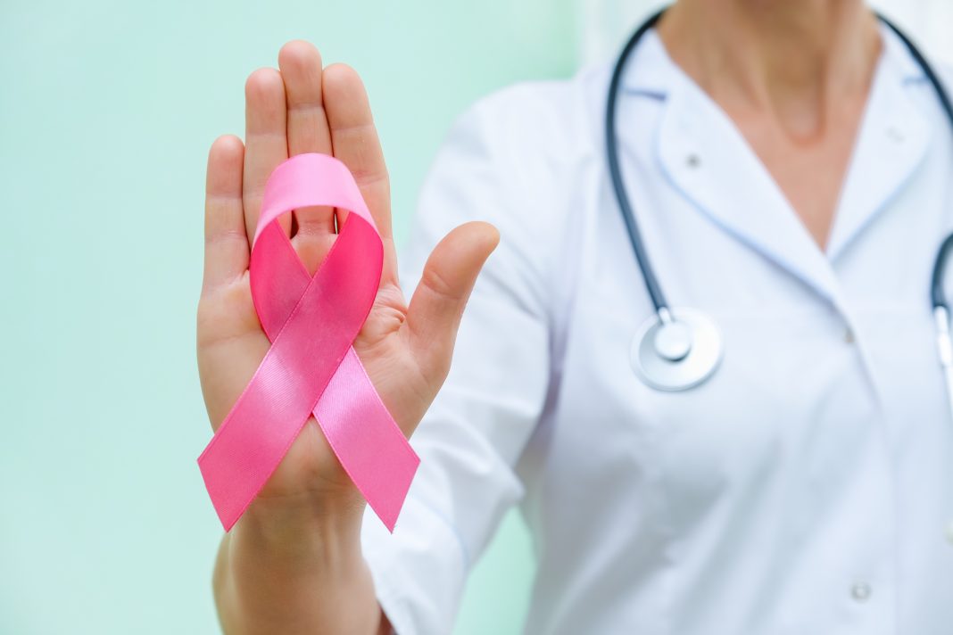 right half of a woman's torso in a doctor's uniform holding up the right hand with a pink breast cancer awareness ribbon on it