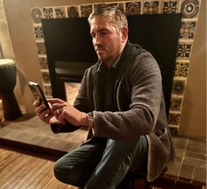 actor Jim Caviezel sitting by a fireplace holding a phone