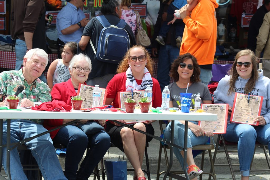 a large crowd behind a white table where three women are sitting with drinks on it.