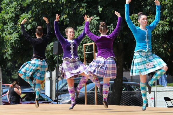 four girls in modern kilts doing a Scottish dance on a wood stage