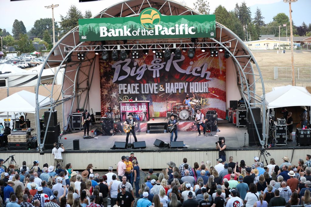 Big & Rich performing on an outdoor stage with the words 'Bank of the Pacific' above it and their name on a banner behind them