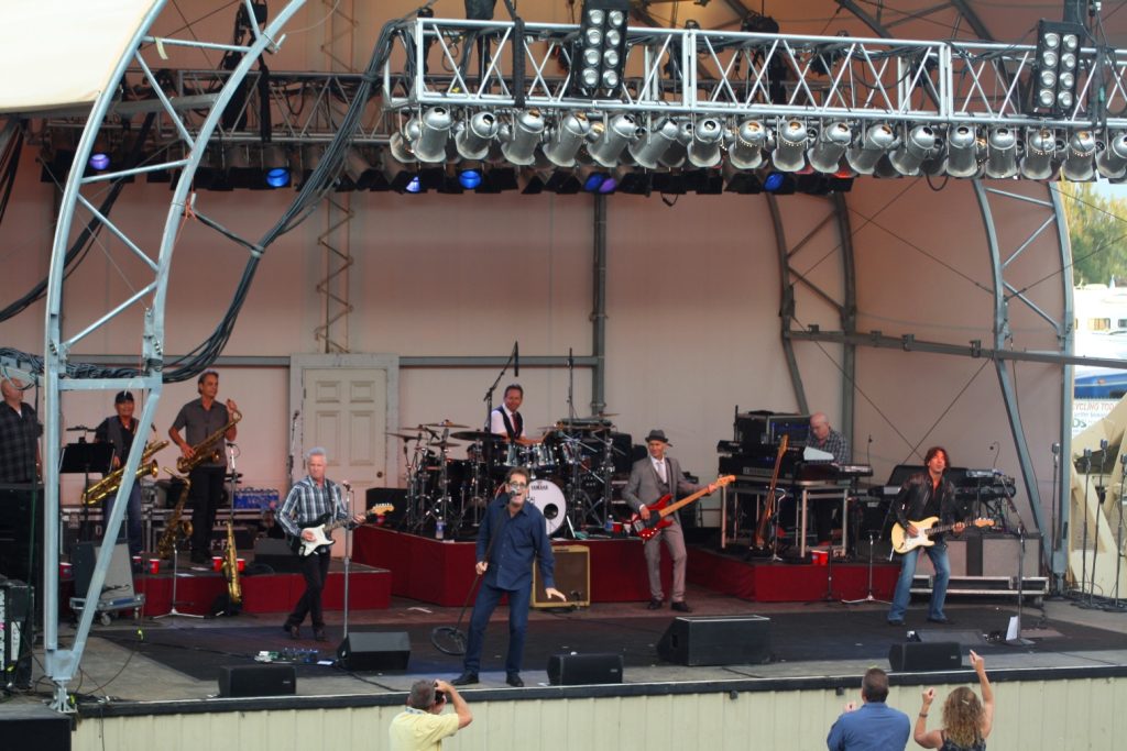 Huey Lewis performing with his band on an outdoor stage with the words 'Bank of the Pacific' above it