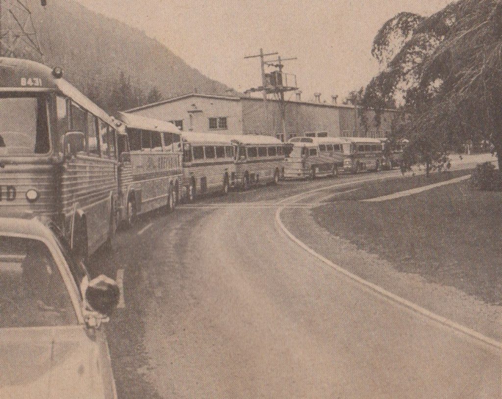 black and white photo of a line of buses backed up on a road with a building in the background.