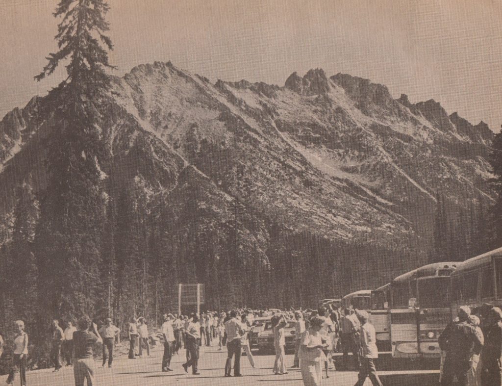 a line of buses with their doors open on North Cascades Highway with mountains in the background. A large number of people are milling around in the street