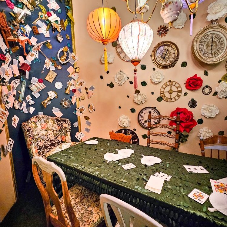 a long table with a green clothe and mismatched chairs in a room with walls covered with different things - flowers, clock faces, playing cards, etc.