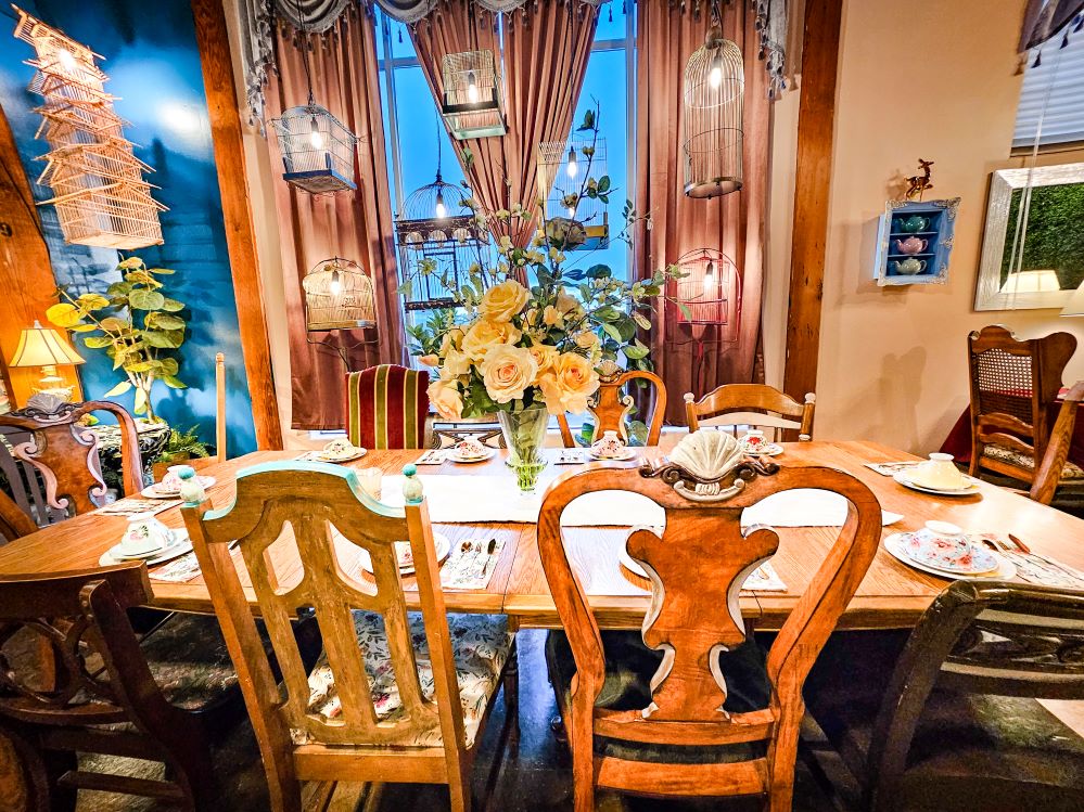 a large wood table with mismatched chairs, set for tea with cups and saucers. Bird cages with lights hand from the ceiling.