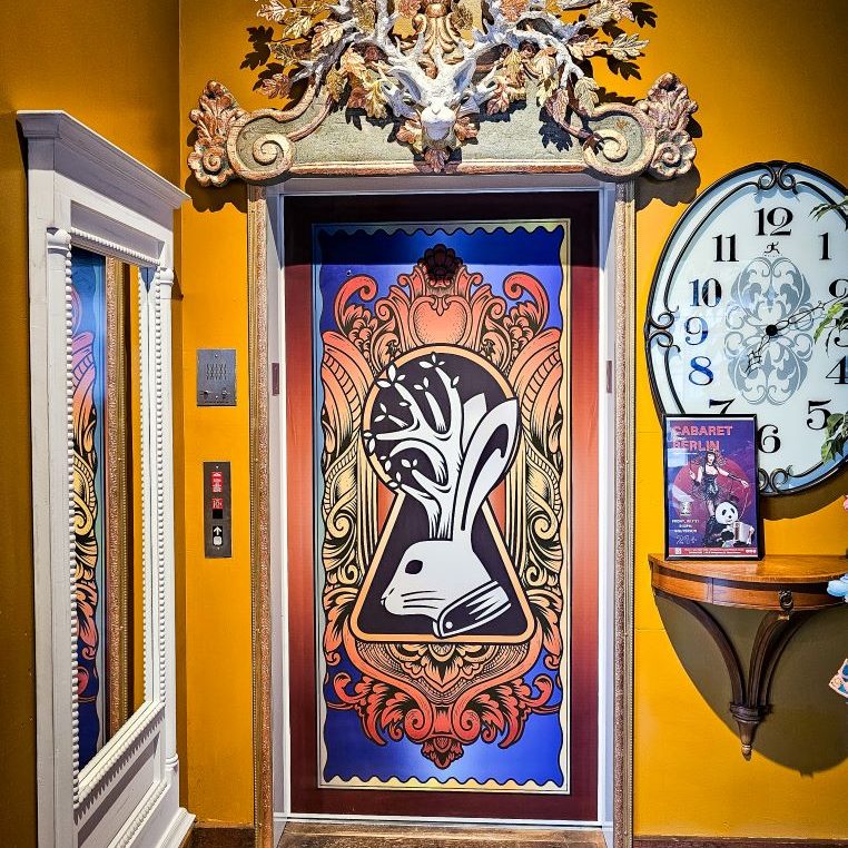 brightly colored elevator door with a blue and orange design including a white rabbit with antlers in the center of a giant keyhole with a black background. A white rabbit head with antlers is above the door, a large clock face is next to it