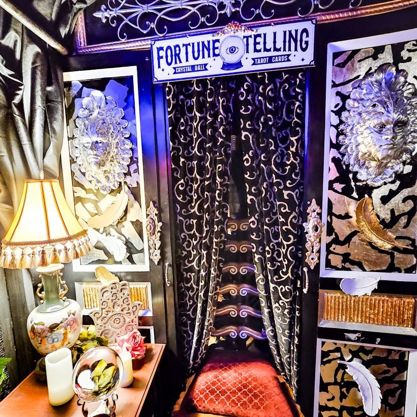 A curtained doorway with a chair in it, lost of oddities on either side and a sign above it that says, 'Fortune Telling. Crystal Ball. Tarot Cards' with a drawing of a crystal ball with an eye in it.