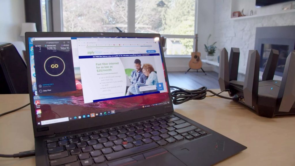 A laptop sitting on a desk with a larger internet router next to it, a guitar and fireplace out of focus in the background