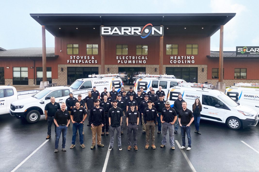 Barron Heating AC Electrical & Plumbing team in black shirts standing in front of the Barron building and several Barron work trucks and vans