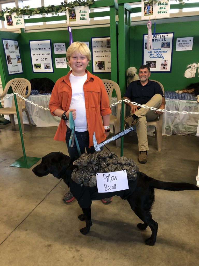 Patrick Swan standing next to his dog wearing a rock costume with a sign on it that says 'pillow basalt'