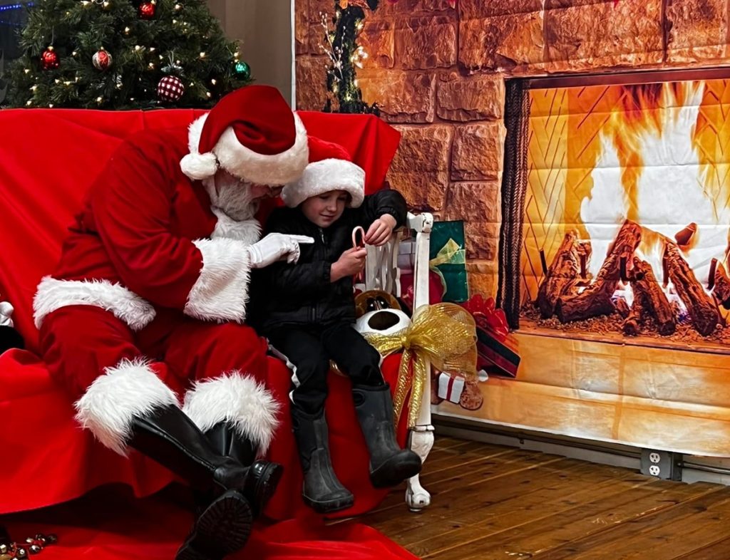 Santa with a child both sitting in a big chair by a photo of a fireplace