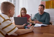 a girl sits at a table on a laptop, a man is next her holding a cellphone and across the table is a young boy holding a tablet