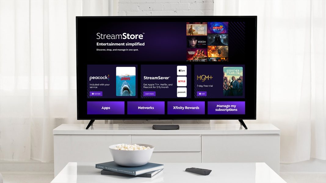 a bowl of popcorn sits on a couple books on a white coffee table with a remote next to it. Behind it it a white TV stand with a large slat screen TV with the Comcast StreamStore App on it. A sound bar is in front of it