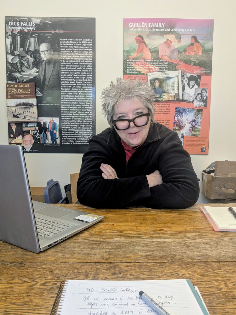 Jo Wolfe, Skagit County Historical Museum Director sitting at a desk with her arms folded. a laptop is open next to her and there is a pen and paper on the desk as well