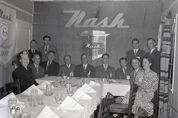 black and white photos showing A group of dressed-up people sit at a T-shaped table with elegant place settings. The word 'Nash" is on the wall behind them.
