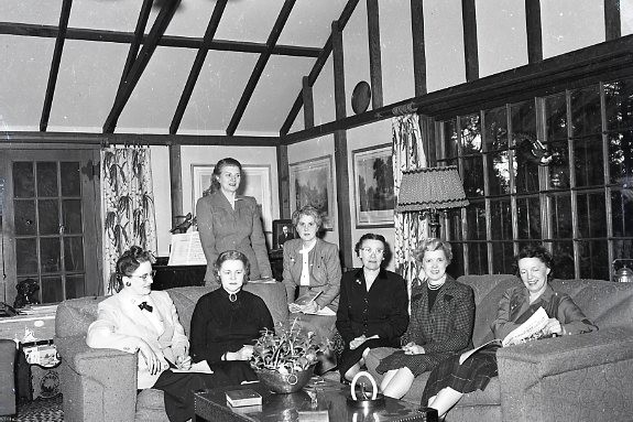 black and white photo of seven woman, all sitting on couches, but one who is standing behind a couch and another who is sitting on a stool.