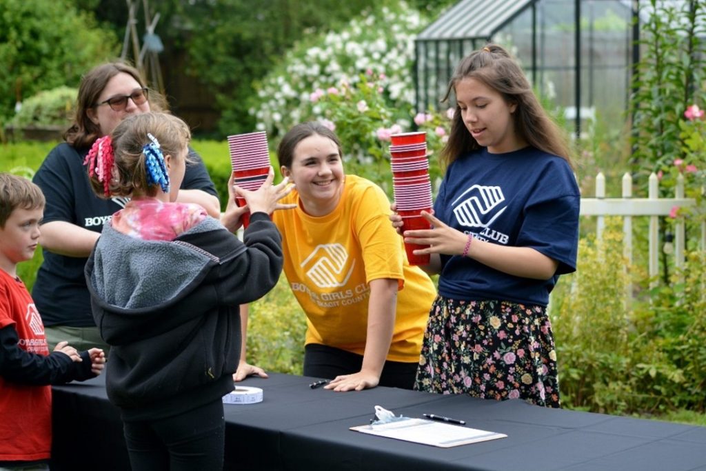 Two girls in Boys & Girls Clubs t-shirts are standing by a table, one is holding stacks of red plastic cups. A woman stands at the end of the table. Two kids are in front of the table, one is holding a stack of cups as well.