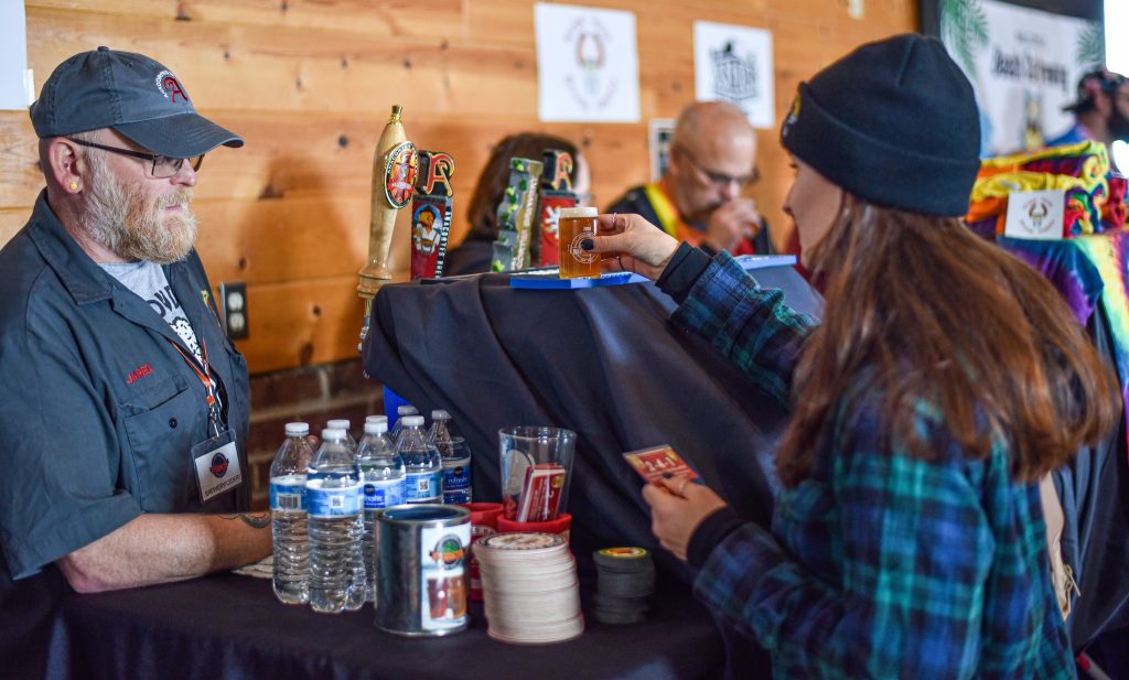 A man serves drinks behind a bar with bottled water next to it. A woman is picking up a shot glass off the top of a beer tap