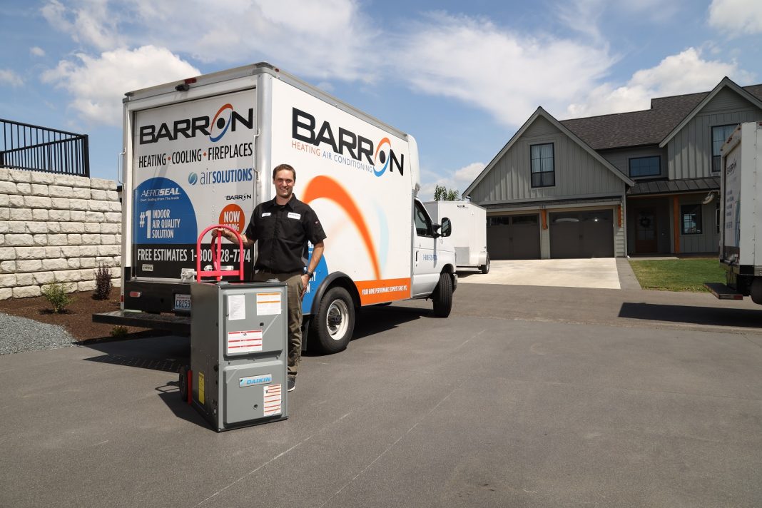 a Barron Heating, AC, Electrical & Plumbing technician standing but a Barron struck with an HVAC on a hand truck at outside a house